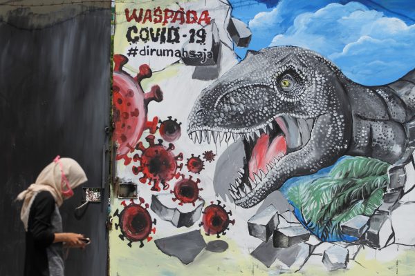 A woman wearing a protective mask walks past a dinosaur depicted on a wall, amid the coronavirus disease (COVID-19) outbreak, in Depok, near Jakarta, Indonesia, 30 March 2020 (Antara Foto/Asprilla Dwi Adha via Reuters).