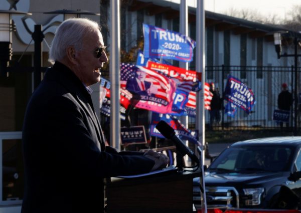 Former vice president Joe Biden speaks as supporters of Donald Trump hold up flags and posters nearby in St Paul, Minnesota, 30 October 2020 (Photo: Reuters/Brian Snyder).
