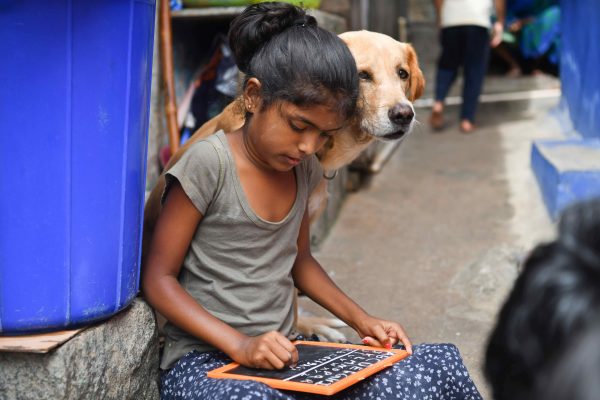 A dog sits next to a girl who is practicing the alphabet on a slate in a slum as schools are shut due the on-going pandemic, Bengaluru, India, 19 Jun 2021 (Photo: Meghana Sastry/SOPA Images/Sipa USA)