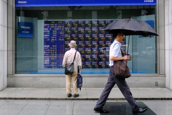 A man holding an umbrella walks in front of an electric board showing Nikkei index a brokerage in Tokyo, Japan, 13 May 2022 (Photo: James Matsumoto/SOPA Images/Sipa USA via Reuters Connect)